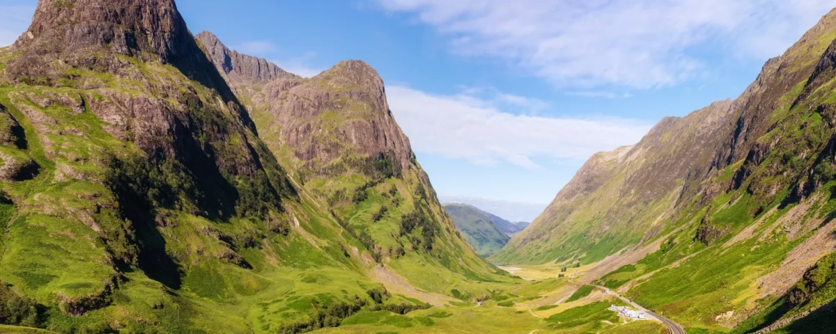 Fotografía panorámica del Valle de Glencoe bajo un cielo despejado y azul. Las montañas rocosas lucen tonos verde esmeralda y ocre iluminados por el sol, transmitiendo la inmensidad y quietud de las Tierras Altas.