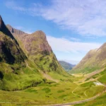 Fotografía panorámica del Valle de Glencoe bajo un cielo despejado y azul. Las montañas rocosas lucen tonos verde esmeralda y ocre iluminados por el sol, transmitiendo la inmensidad y quietud de las Tierras Altas.