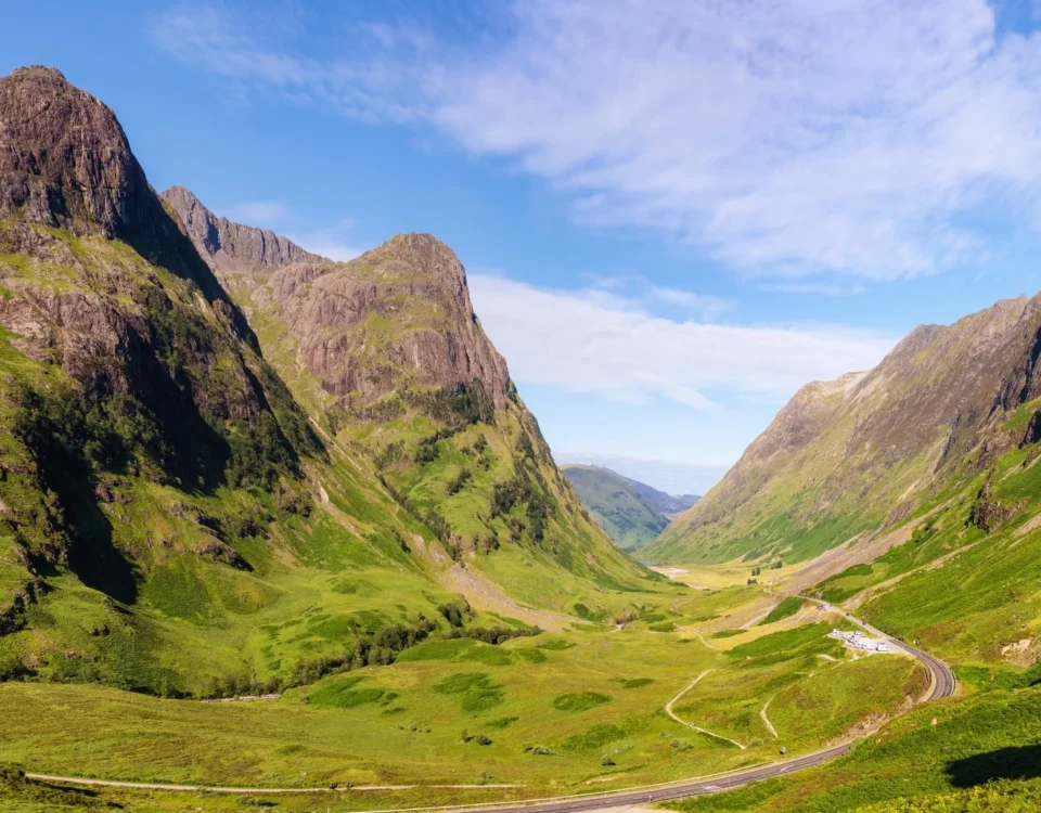 Fotografía panorámica del Valle de Glencoe bajo un cielo despejado y azul. Las montañas rocosas lucen tonos verde esmeralda y ocre iluminados por el sol, transmitiendo la inmensidad y quietud de las Tierras Altas.
