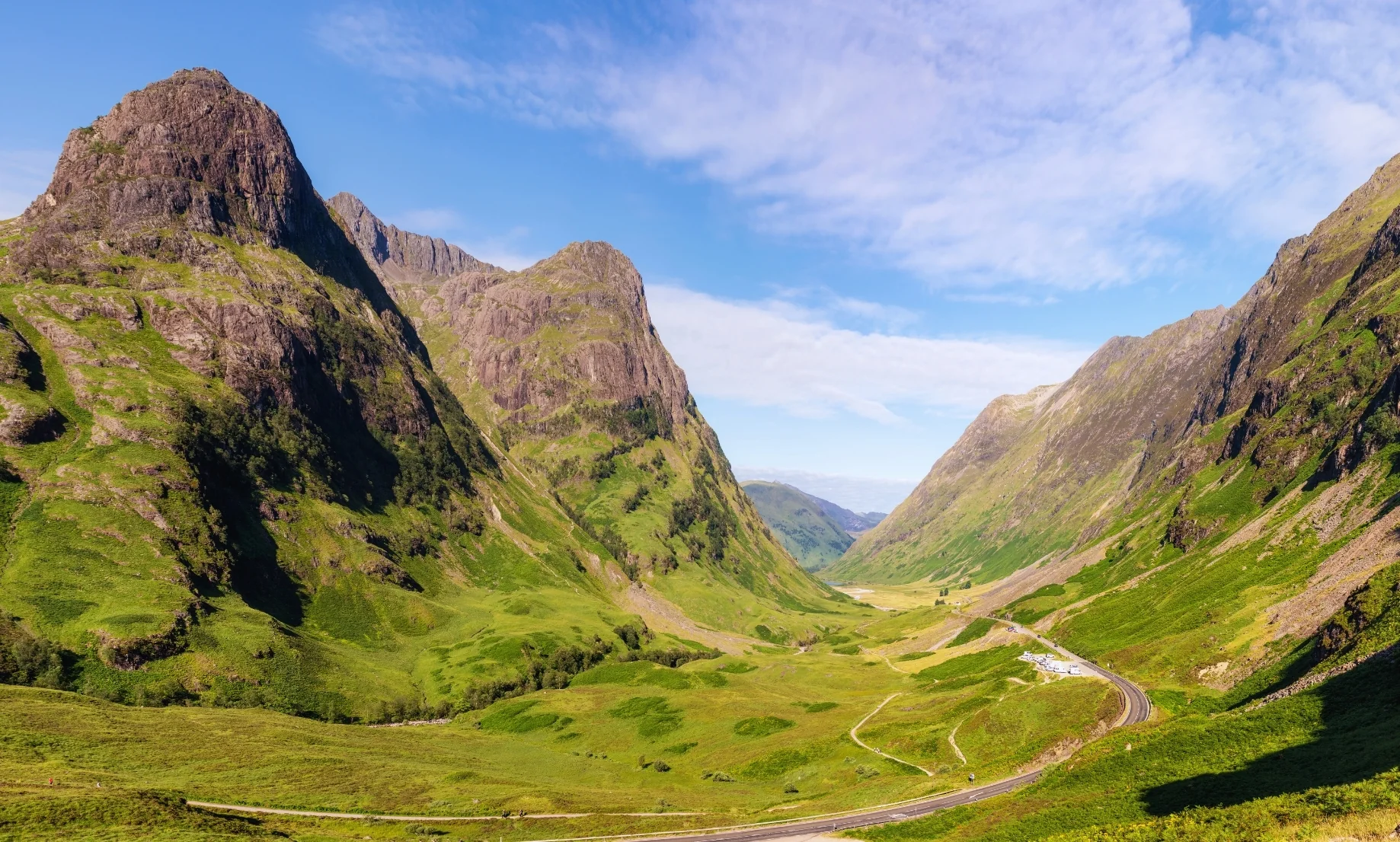 Fotografía panorámica del Valle de Glencoe bajo un cielo despejado y azul. Las montañas rocosas lucen tonos verde esmeralda y ocre iluminados por el sol, transmitiendo la inmensidad y quietud de las Tierras Altas.