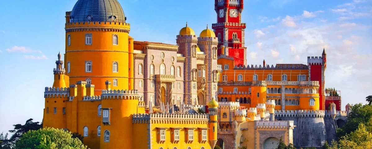 Fotografía panorámica elevada del Palacio da Pena en Sintra, Portugal, bajo un cielo azul con nubes ligeras. Se aprecian las distintivas torres de colores amarillo brillante y rojo intenso rodeadas por la exuberante vegetación verde del parque.