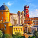 Fotografía panorámica elevada del Palacio da Pena en Sintra, Portugal, bajo un cielo azul con nubes ligeras. Se aprecian las distintivas torres de colores amarillo brillante y rojo intenso rodeadas por la exuberante vegetación verde del parque.