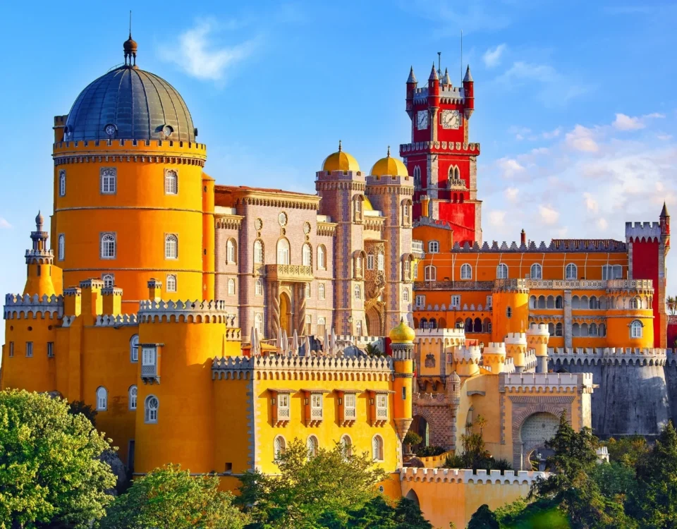 Fotografía panorámica elevada del Palacio da Pena en Sintra, Portugal, bajo un cielo azul con nubes ligeras. Se aprecian las distintivas torres de colores amarillo brillante y rojo intenso rodeadas por la exuberante vegetación verde del parque.