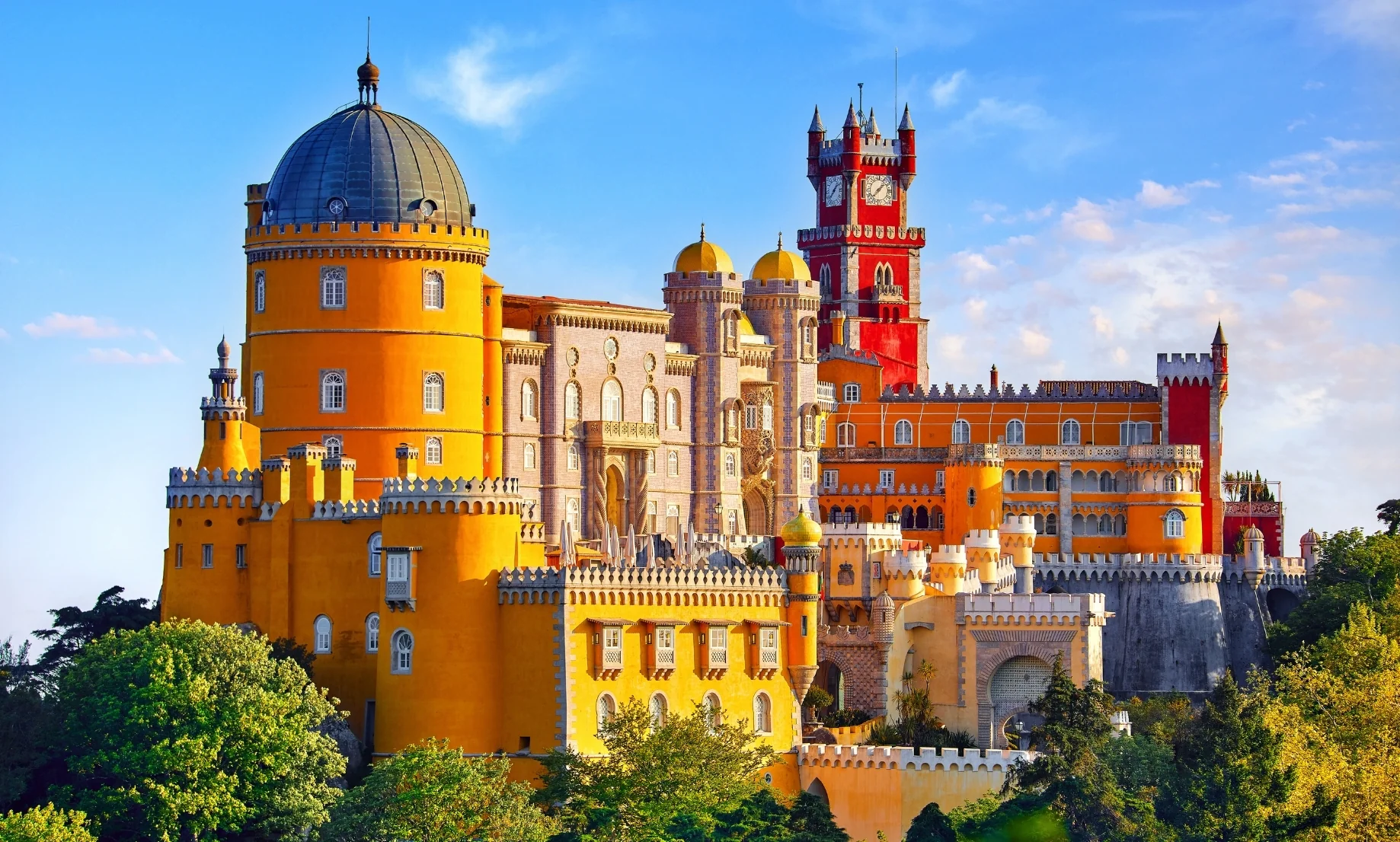 Fotografía panorámica elevada del Palacio da Pena en Sintra, Portugal, bajo un cielo azul con nubes ligeras. Se aprecian las distintivas torres de colores amarillo brillante y rojo intenso rodeadas por la exuberante vegetación verde del parque.