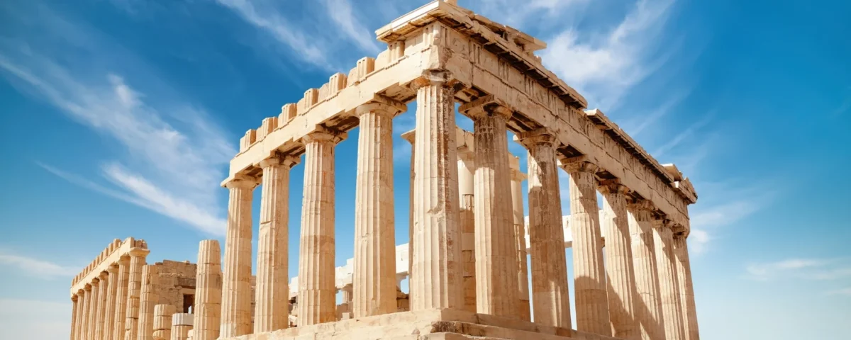 Fotografía panorámica a nivel de suelo del templo del Partenón en la Acrópolis de Atenas, Grecia, mostrando sus imponentes columnas dóricas de mármol contra un cielo azul vibrante cubierto de nubes blancas difusas. En primer plano, bloques de mármol antiguos dispersos.
