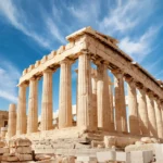 Fotografía panorámica a nivel de suelo del templo del Partenón en la Acrópolis de Atenas, Grecia, mostrando sus imponentes columnas dóricas de mármol contra un cielo azul vibrante cubierto de nubes blancas difusas. En primer plano, bloques de mármol antiguos dispersos.