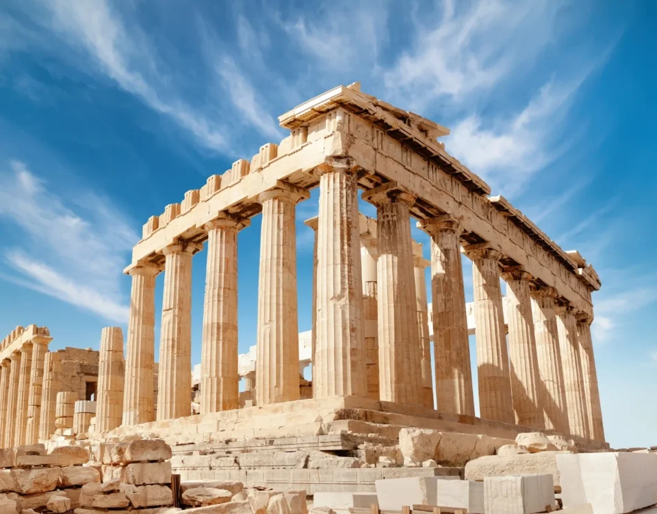 Fotografía panorámica a nivel de suelo del templo del Partenón en la Acrópolis de Atenas, Grecia, mostrando sus imponentes columnas dóricas de mármol contra un cielo azul vibrante cubierto de nubes blancas difusas. En primer plano, bloques de mármol antiguos dispersos.