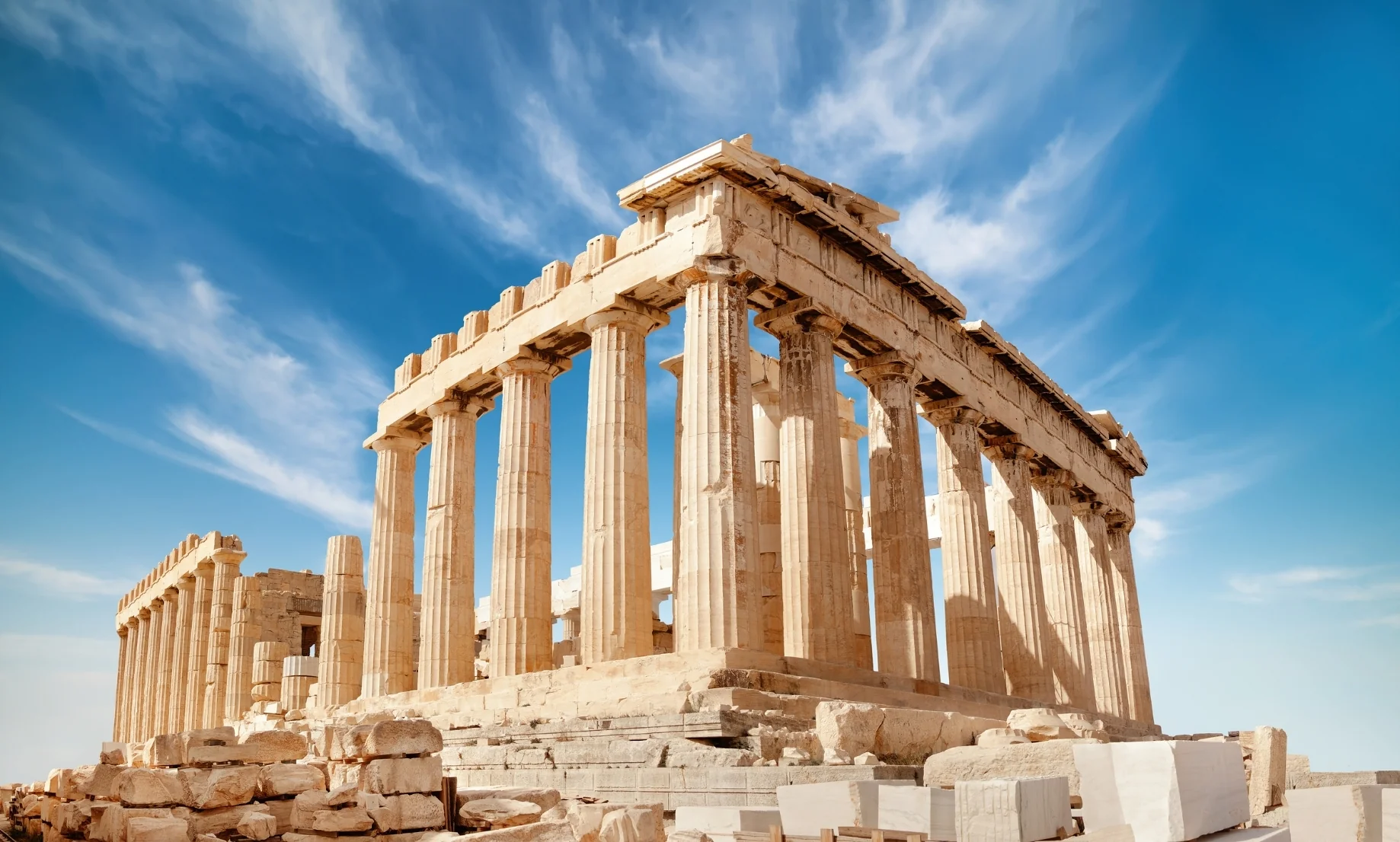 Fotografía panorámica a nivel de suelo del templo del Partenón en la Acrópolis de Atenas, Grecia, mostrando sus imponentes columnas dóricas de mármol contra un cielo azul vibrante cubierto de nubes blancas difusas. En primer plano, bloques de mármol antiguos dispersos.