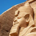 Primer plano de la cabeza de la estatua colosal del faraón Ramsés II tallada en roca en Abu Simbel, Egipto, destacando la textura rugosa de la arenisca bajo un cielo azul despejado.