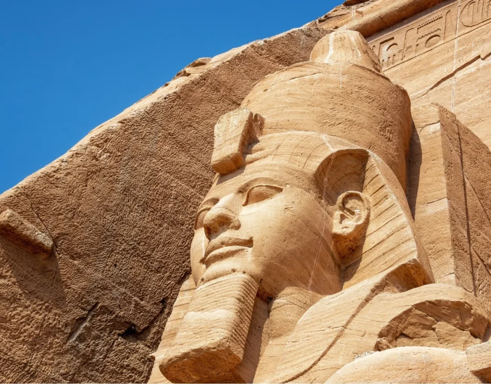 Primer plano de la cabeza de la estatua colosal del faraón Ramsés II tallada en roca en Abu Simbel, Egipto, destacando la textura rugosa de la arenisca bajo un cielo azul despejado.