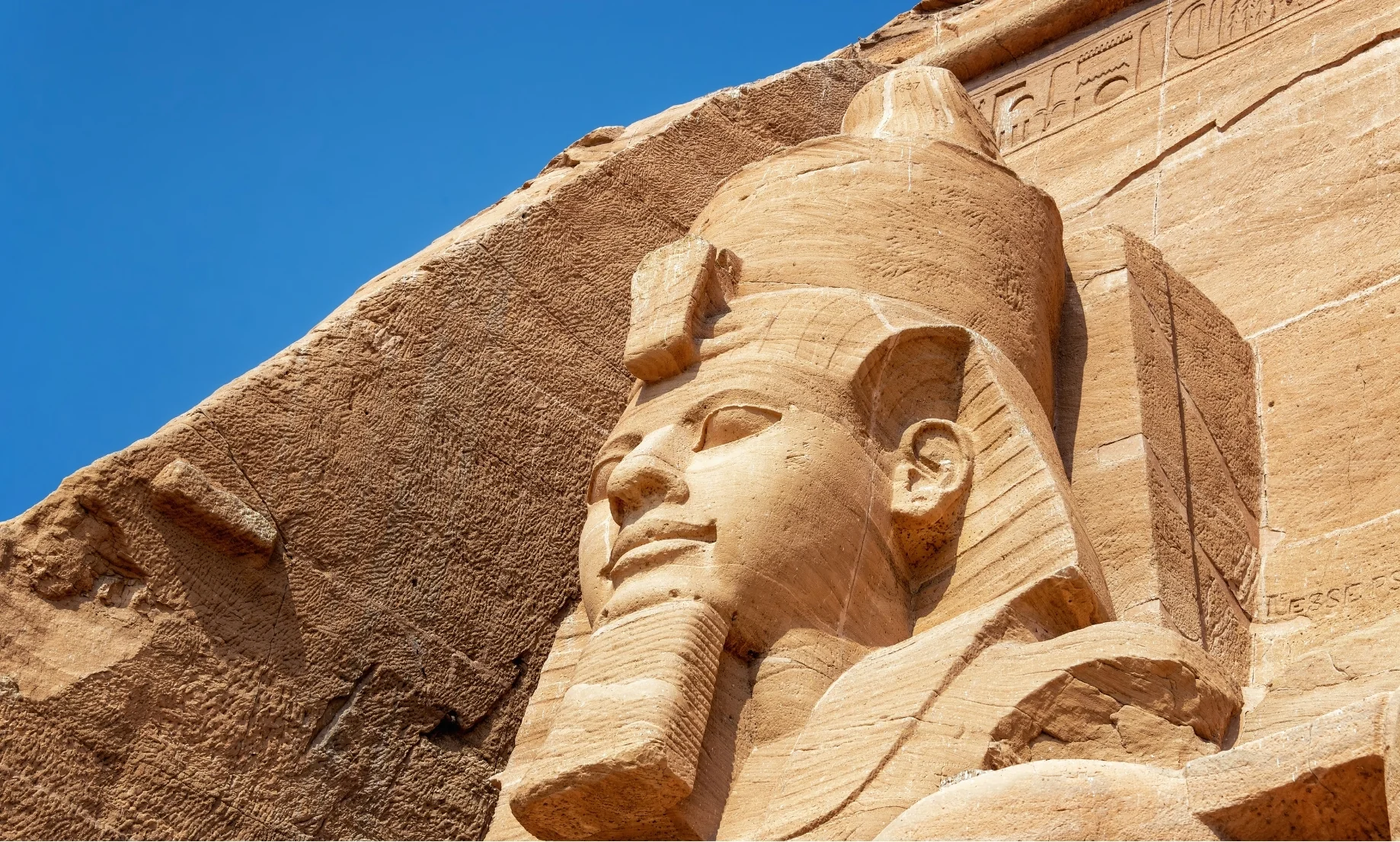 Primer plano de la cabeza de la estatua colosal del faraón Ramsés II tallada en roca en Abu Simbel, Egipto, destacando la textura rugosa de la arenisca bajo un cielo azul despejado.