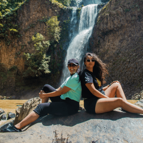 Dos amigas excursionistas sentadas sonrientes espalda con espalda sobre una gran roca con vistas a una alta cascada que cae en una piscina de agua dorada en un cañón de roca.