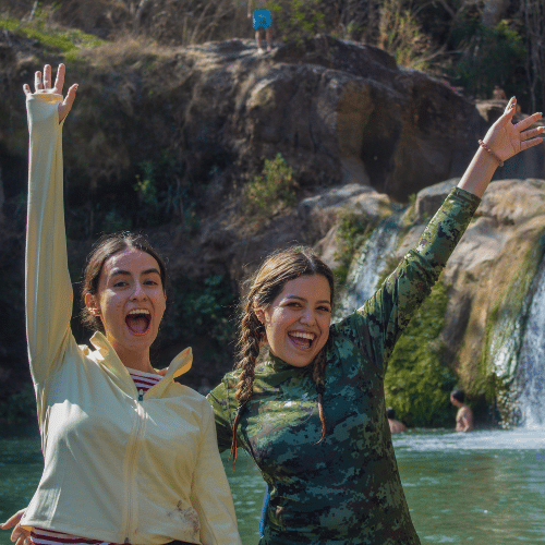 Dos jóvenes sonriendo y celebrando con los brazos en alto frente a una cascada y pozas de agua natural en Comala.