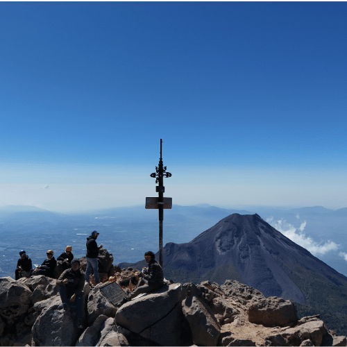 Grupo de excursionistas descansando en la cumbre rocosa del Nevado de Colima, contemplando el Volcán de Fuego bajo un cielo completamente despejado.