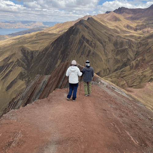 Pareja de excursionistas abrigados caminando por la cresta afilada y colorida de la montaña Pallay Punchu, con un valle profundo y una laguna al fondo.