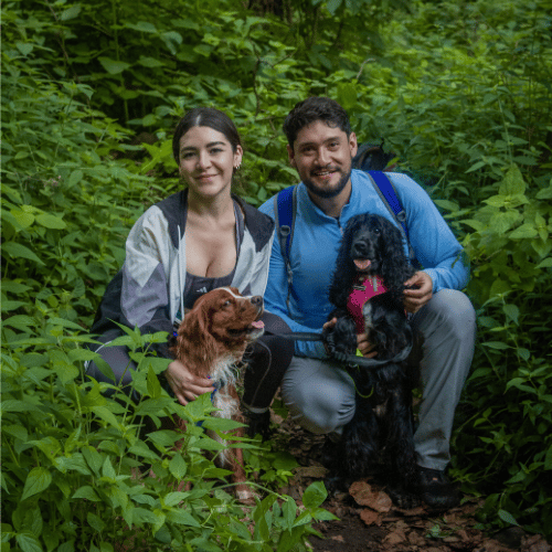Pareja de excursionistas sonriendo mientras abrazan a sus dos perros en un sendero rodeado de denso follaje verde.