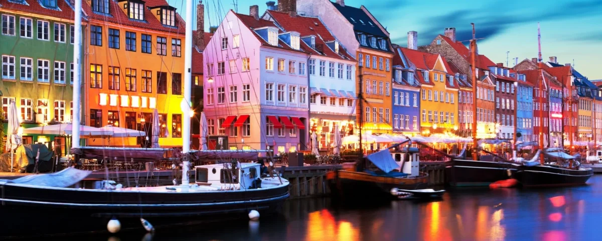 Vista del puerto histórico de Nyhavn en Dinamarca durante la hora azul. Las luces de los edificios mercantiles y restaurantes pintan el agua oscura del canal junto a antiguos veleros.