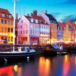 Vista del puerto histórico de Nyhavn en Dinamarca durante la hora azul. Las luces de los edificios mercantiles y restaurantes pintan el agua oscura del canal junto a antiguos veleros.