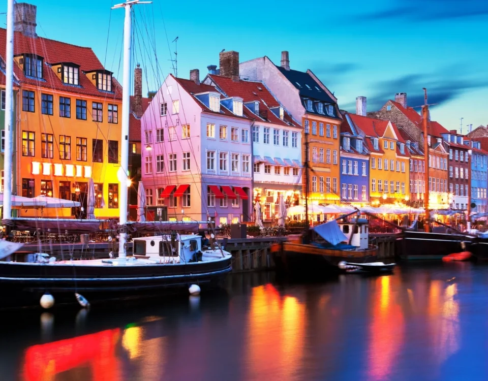 Vista del puerto histórico de Nyhavn en Dinamarca durante la hora azul. Las luces de los edificios mercantiles y restaurantes pintan el agua oscura del canal junto a antiguos veleros.