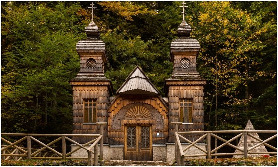Vista frontal de la histórica Capilla Rusa (Ruska kapelica) de madera en Eslovenia, con sus dos torres gemelas ornamentadas y tallados folclóricos, situada en un denso bosque de otoño cerca del paso Vršič.