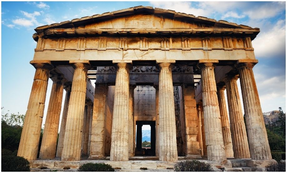 Fotografía frontal del Templo de Hefesto en Atenas, destacando sus robustas columnas dóricas de mármol teñidas de tonos cálidos por el sol, bajo un cielo azul con nubes ligeras.
