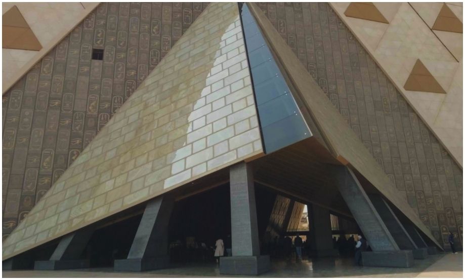 Toma frontal de la colosal entrada del Gran Museo Egipcio en Giza, que se destaca por su estructura piramidal de vidrio y metal frente a una inmensa pared cubierta de miles de jeroglíficos antiguos y paneles triangulares. Se aprecian personas a la distancia para mostrar la escala monumental.