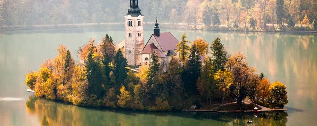Fotografía de la isla central del Lago de Bled en Eslovenia, donde una iglesia histórica se alza entre árboles con denso follaje amarillo y naranja de otoño, reflejándose perfectamente en el agua verde y serena.
