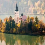 Fotografía de la isla central del Lago de Bled en Eslovenia, donde una iglesia histórica se alza entre árboles con denso follaje amarillo y naranja de otoño, reflejándose perfectamente en el agua verde y serena.