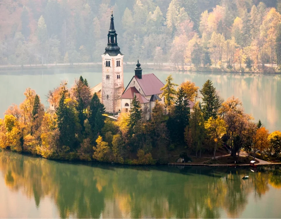 Fotografía de la isla central del Lago de Bled en Eslovenia, donde una iglesia histórica se alza entre árboles con denso follaje amarillo y naranja de otoño, reflejándose perfectamente en el agua verde y serena.