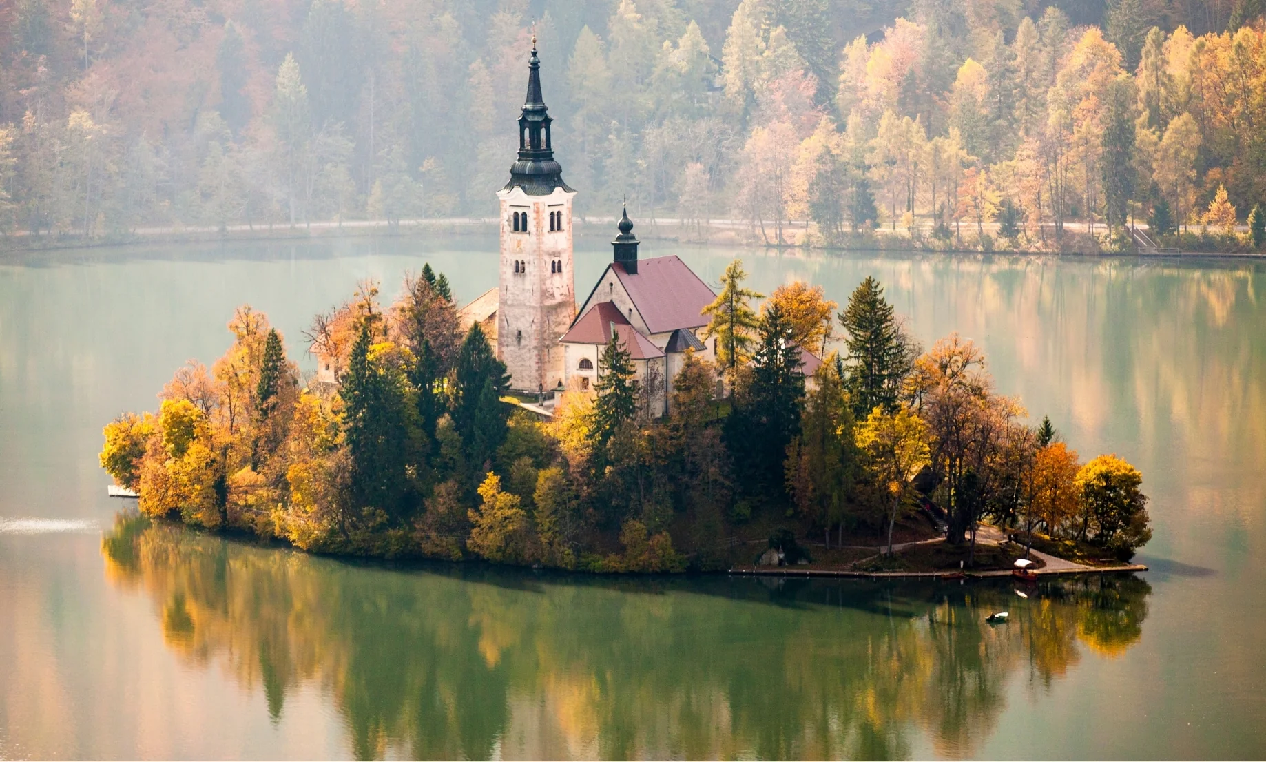 Fotografía de la isla central del Lago de Bled en Eslovenia, donde una iglesia histórica se alza entre árboles con denso follaje amarillo y naranja de otoño, reflejándose perfectamente en el agua verde y serena.