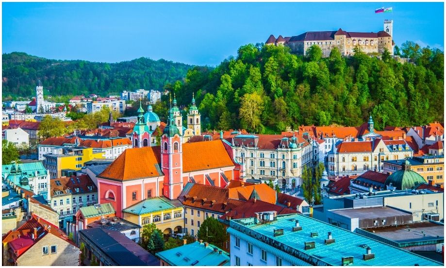 Vista aérea del vibrante centro histórico de Liubliana en Eslovenia, destacando una iglesia de color salmón y el castillo medieval en la cima de una colina verde bajo un cielo azul brillante.
