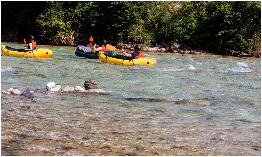 Grupo de aventureros haciendo rafting en balsas inflables amarillas sobre las aguas poco profundas y cristalinas del río Soča en Bovec, rodeados de bosque y rocas de río.