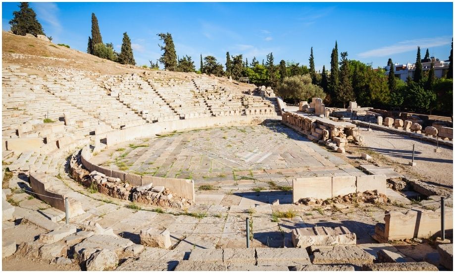 Vista amplia del Teatro de Dionisio en Atenas, mostrando sus antiguas gradas de piedra semicirculares y el escenario de mármol rodeado de árboles verdes bajo un cielo azul despejado.