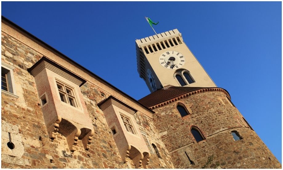 Fotografía arquitectónica de la fortaleza del Castillo de Ljubljana, mostrando el contraste entre la mampostería medieval, las ventanas clásicas y la esfera blanca del reloj iluminada por el sol.